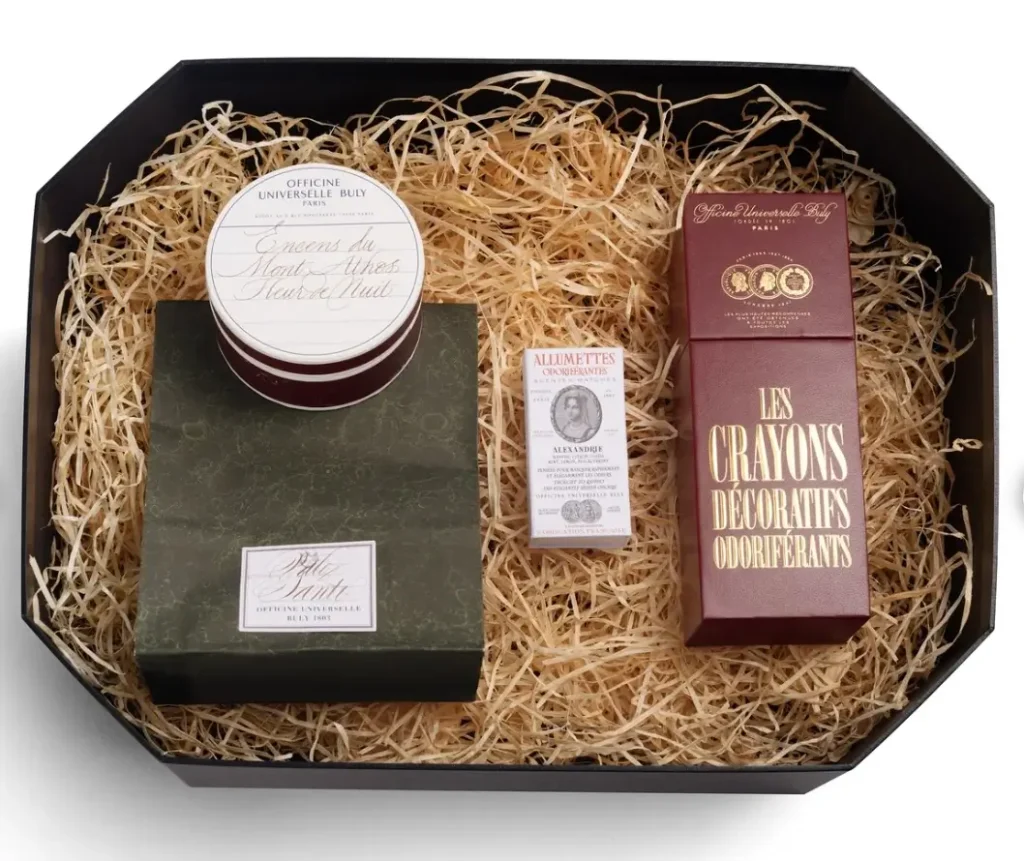 Octagonal gift box filled with shredded straw, presenting Officine Universelle Buly’s scented interior set with a tin of Mount Athos incense, a green stone incense holder, a box of scented matches, and a burgundy case of decorative scented diffuser pencils