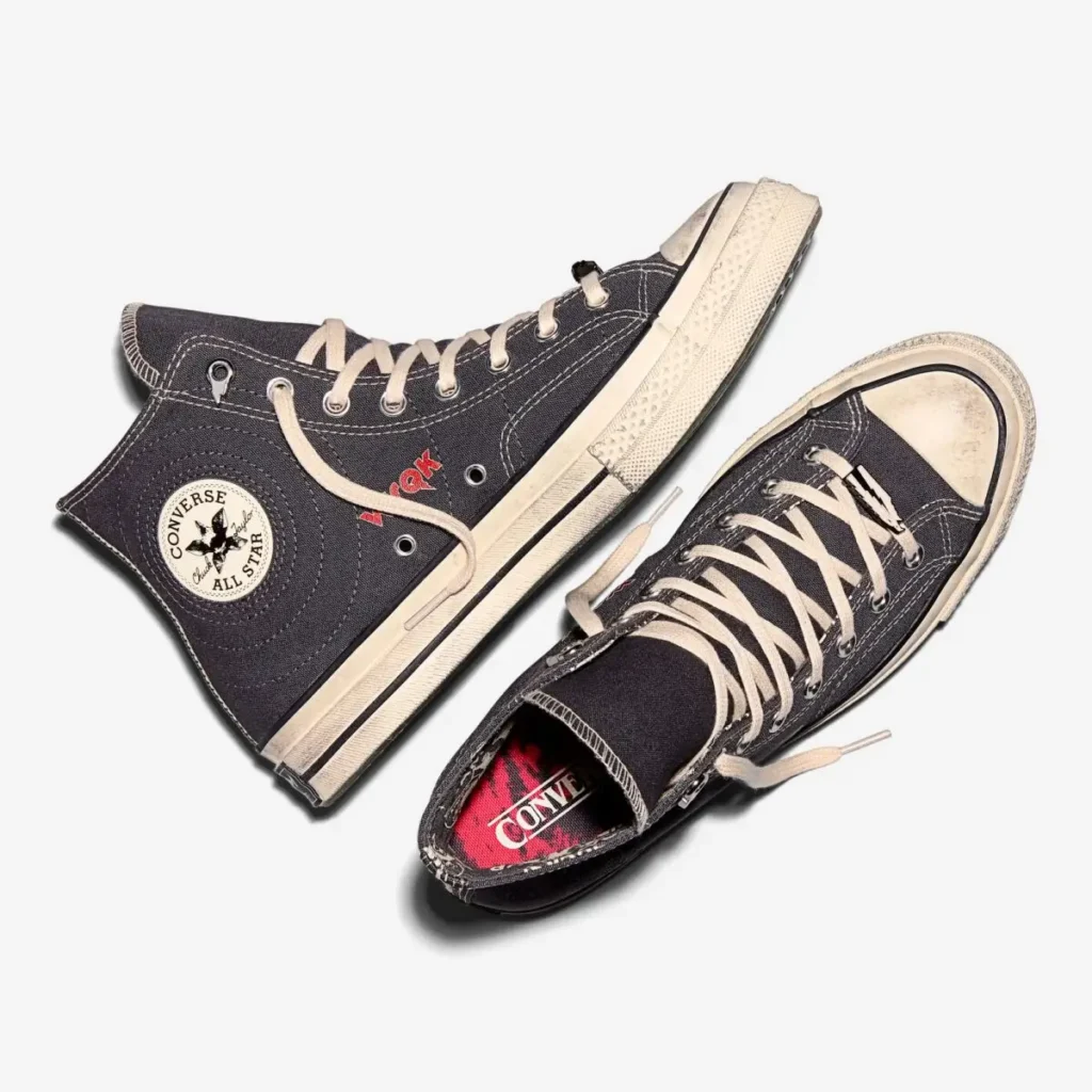 Top-down product view of the Stranger Things x Converse Chuck 70 “WSQK The Squawk” sneakers, showing the charcoal canvas upper, cream laces, aged off-white sole, WSQK red lettering on the side, and a red graphic insole with the Converse logo. One shoe is laid flat while the other rests angled on top against a light background