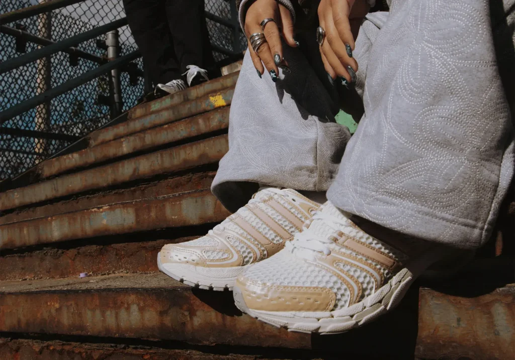 A close-up of a person wearing the adidas Adistar Control 5 sneakers in a cream-and-white colorway while sitting on rusted metal steps, with embroidered grey pants and layered silver rings visible, capturing a street-style running-meets-fashion aesthetic