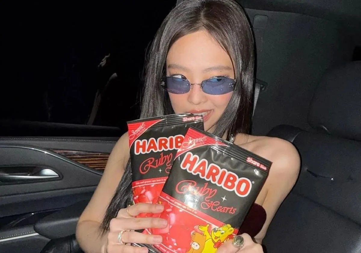 Jennie from BLACKPINK sits in a car wearing slim blue-tinted sunglasses and holding two limited-edition HARIBO Ruby Hearts gummy bags, smiling as she shows the black-and-red packaging to the camera