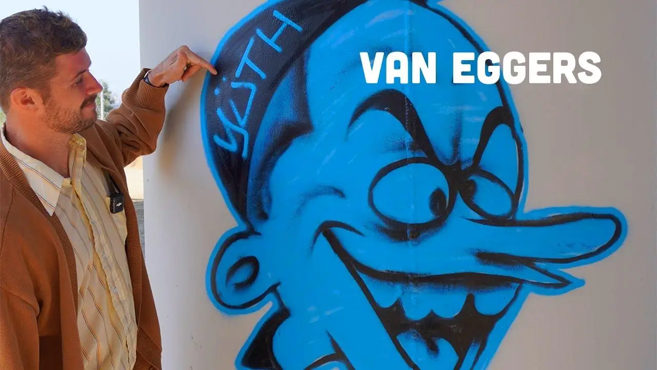 Man pointing at a blue graffiti mural of a cartoon face painted on a wall, with the words “VAN EGGERS” overlaid, showing the artist beside the spray-painted street artwork in daylight