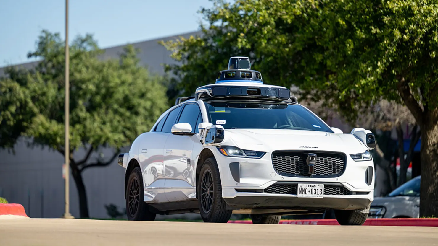 Waymo autonomous Jaguar I-Pace robotaxi driving on a sunny city street with roof-mounted lidar sensors