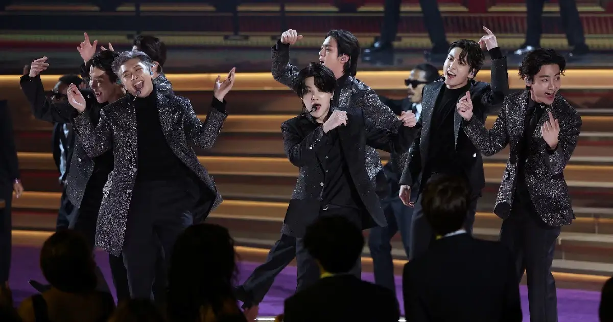 BTS performing live on stage in coordinated black and silver outfits during a concert appearance, with all seven members dancing and singing together as fans watch from the audience, marking the group’s reunion following military servic