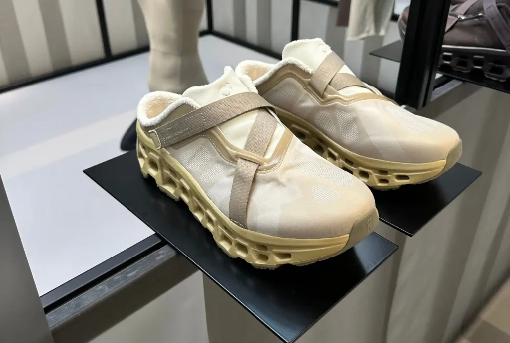 Beige slip-on sneakers displayed on a black platform at Paris Fashion Week, featuring translucent mesh uppers, fleece-lined collars, cross-strap closures, and chunky perforated midsoles, representing On Share’s minimalist performance-driven footwear design