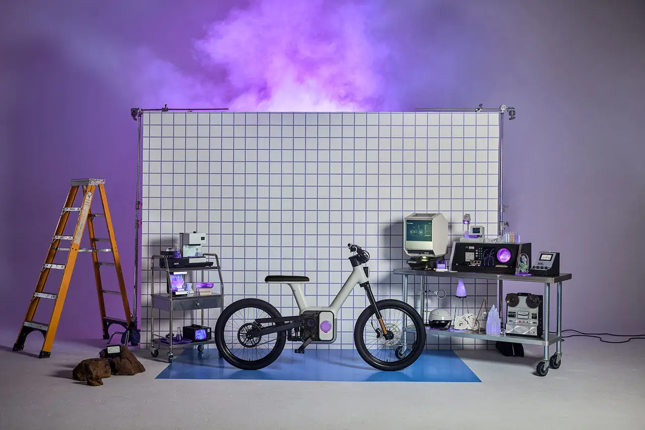 ALSO TM-B electric bike displayed in a studio setting with a modular DreamRide system, surrounded by lab-style equipment, grid backdrop, and purple lighting, emphasizing its software-driven, experimental design ethos