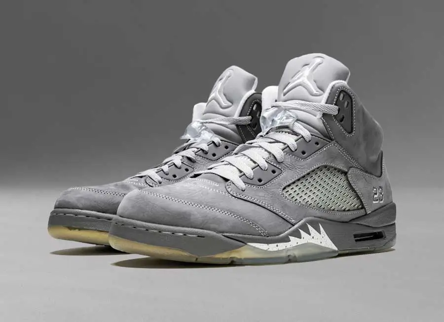 Air Jordan 5 “Wolf Grey” sneakers shown in a three-quarter view, featuring a grey suede upper, reflective silver tongues, mesh side panels, and an icy translucent outsole against a neutral studio background