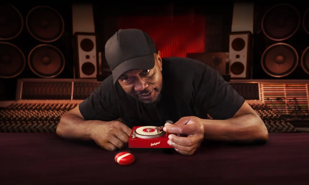 Producer Timbaland leans over a studio mixing desk while holding a tiny Babybel-branded turntable playing a mini vinyl record, with speakers and recording equipment in the background