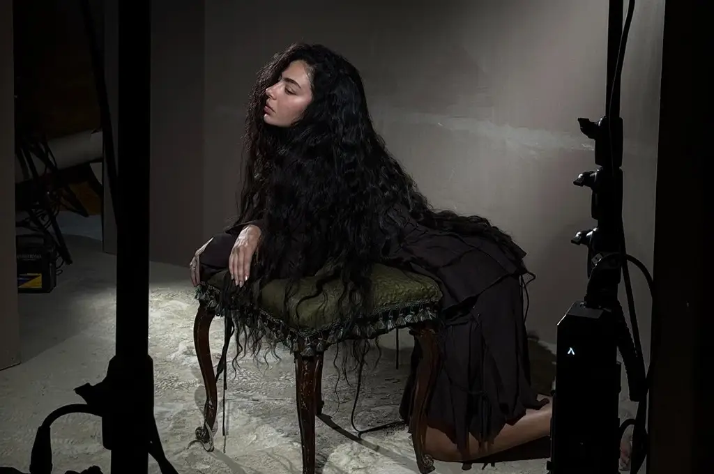 Moody studio portrait of a pop artist Charlie XCX with long dark hair reclining on a vintage chair under dramatic lighting, evoking a dense, atmospheric wall of sound aesthetic