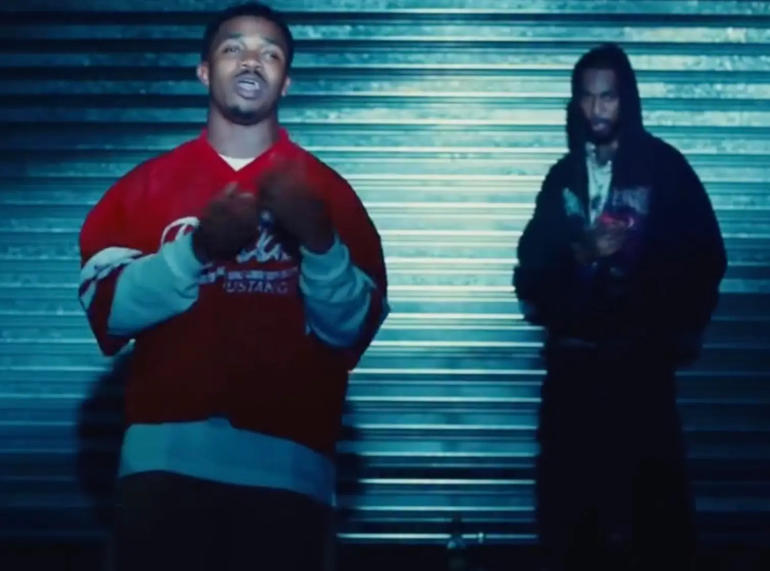 Trap Dickey and Key Glock stand in front of a corrugated metal shutter under cool blue lighting, one in a red jersey gesturing toward the camera while the other, in a dark hoodie, watches from the background