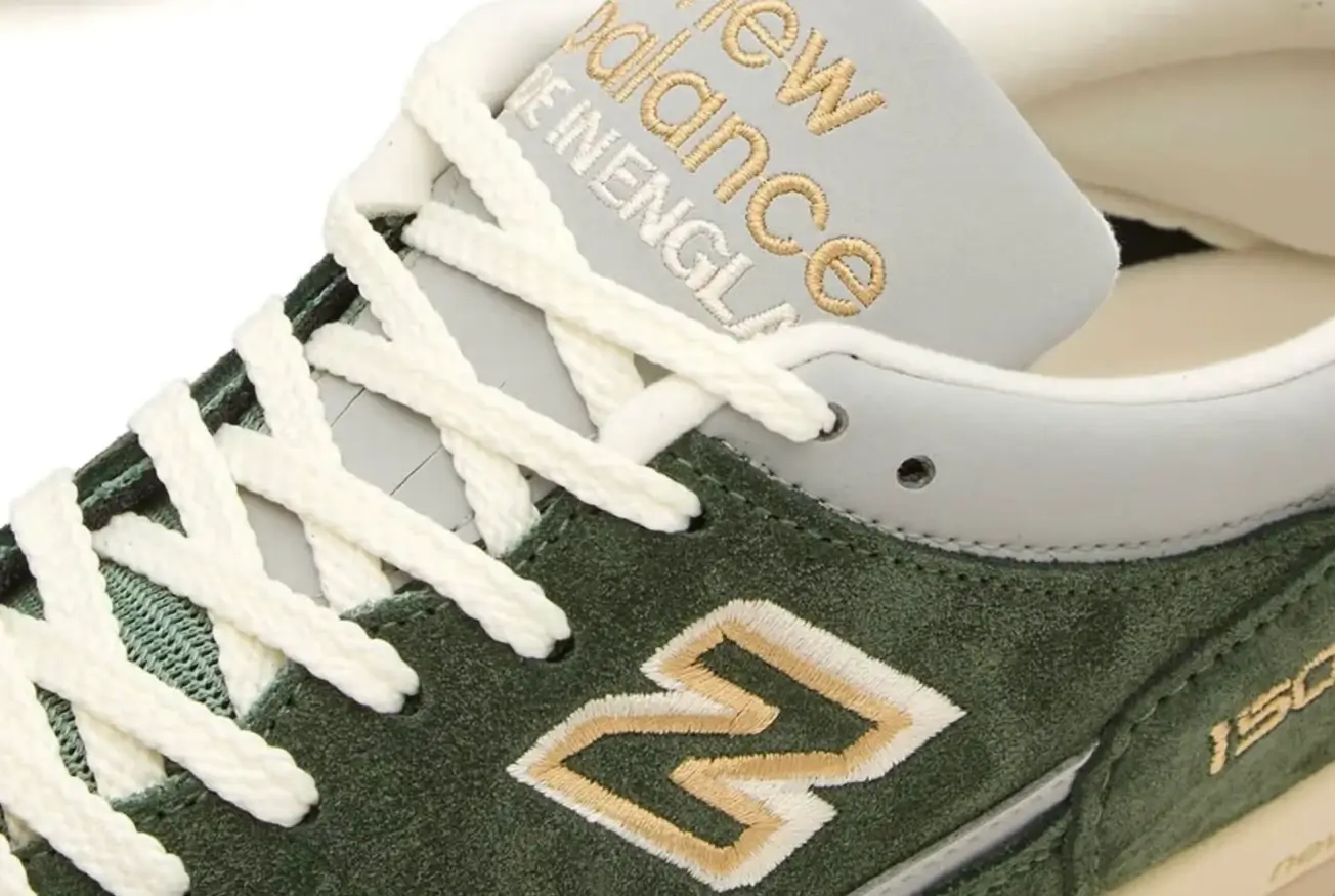Close-up detail of the New Balance 1500 Made in UK “Kombu Green” sneaker, focusing on the deep green suede upper, pale green mesh underlay, white rope laces, gold-and-white embroidered “N” logo, and the grey tongue with gold “new balance ENGLAND” branding, photographed on a white background
