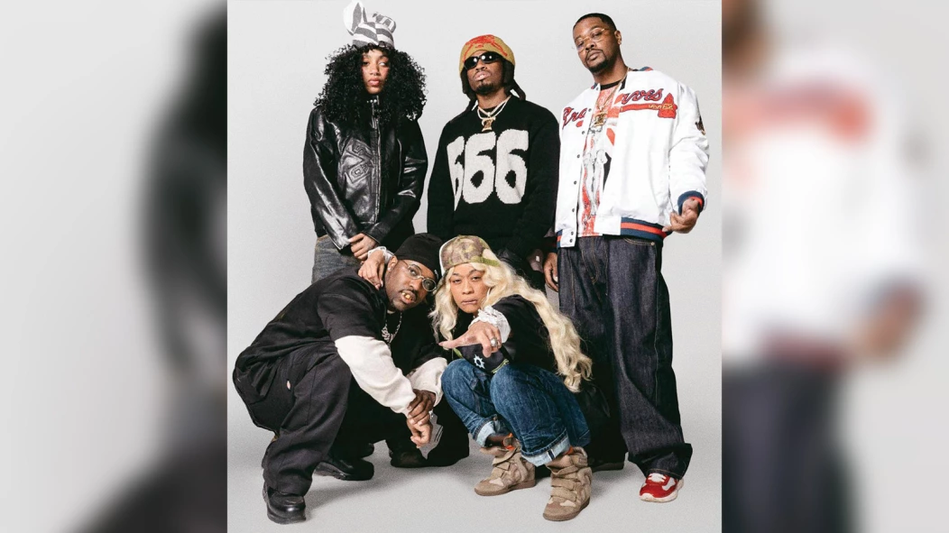 Group portrait of Denzel Curry, BKTHERULA, Laser Dim 700 and collaborators posing in streetwear against a light studio backdrop, wearing leather jackets, graphic knits, baggy jeans, and sneakers in a gritty hip-hop promo shoot