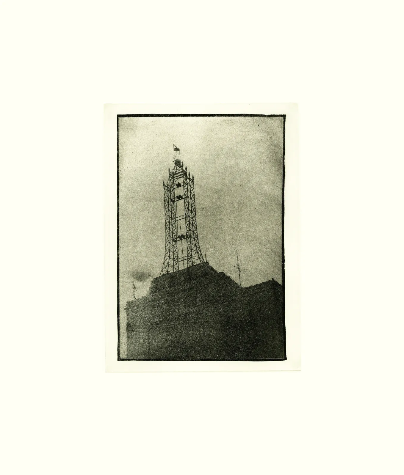 Photogravure-style print showing a skeletal metal radio or transmission tower rising from the roof of a dark, blocky building, framed by a hand-drawn black border against a pale gray, grainy sky