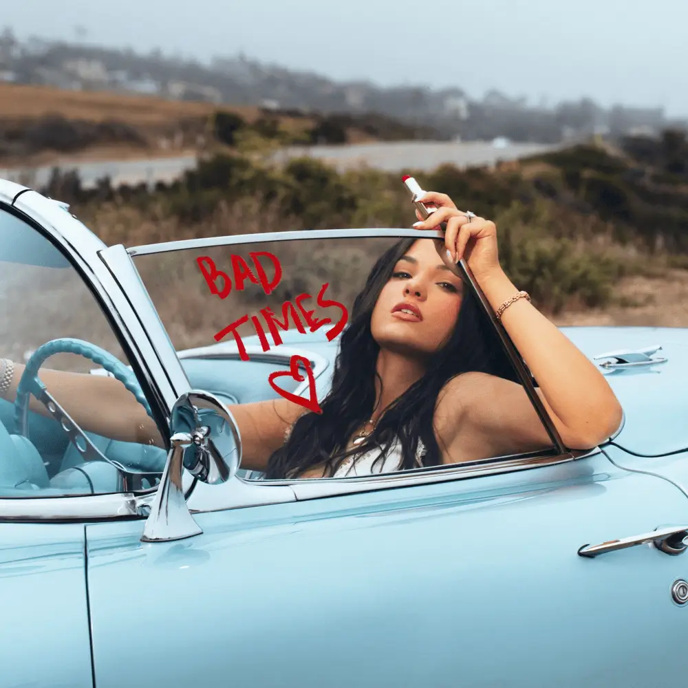 Jessica Baio eaning out of a light blue vintage convertible, holding red lipstick, with the words “BAD TIMES” and a hand-drawn heart written on the car window against a muted countryside backdrop