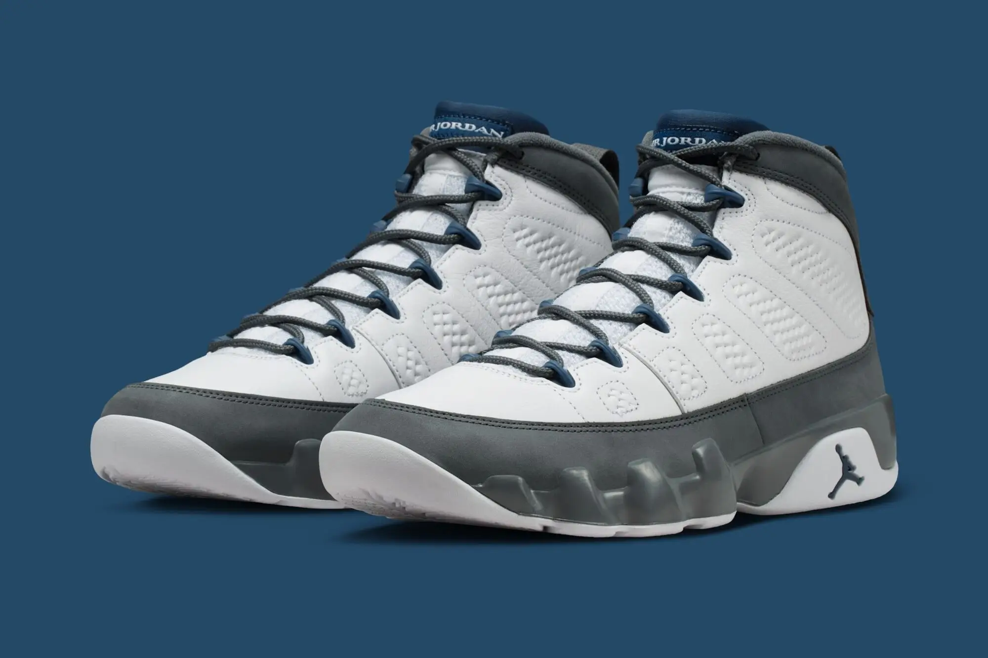 Pair of Air Jordan 9 “Flint Grey” sneakers in white leather with grey mudguard and blue accents on a blue studio background