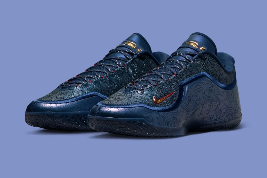 Pair of Nike LeBron 23 “Out For Redemption” basketball shoes in dark navy with embossed graphic patterning, red-and-gold Swoosh, speckled black midsole, and gold LeBron logos on the tongues, photographed against a muted blue background