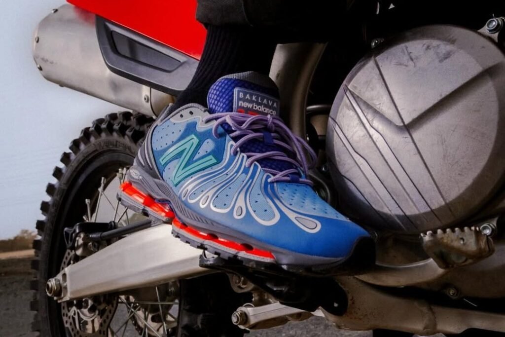 Baklava x New Balance 1890 “Cyborg Tears” sneaker in blue and grey with reflective overlays and purple laces, worn on foot resting against a motorcycle engine and rear wheel