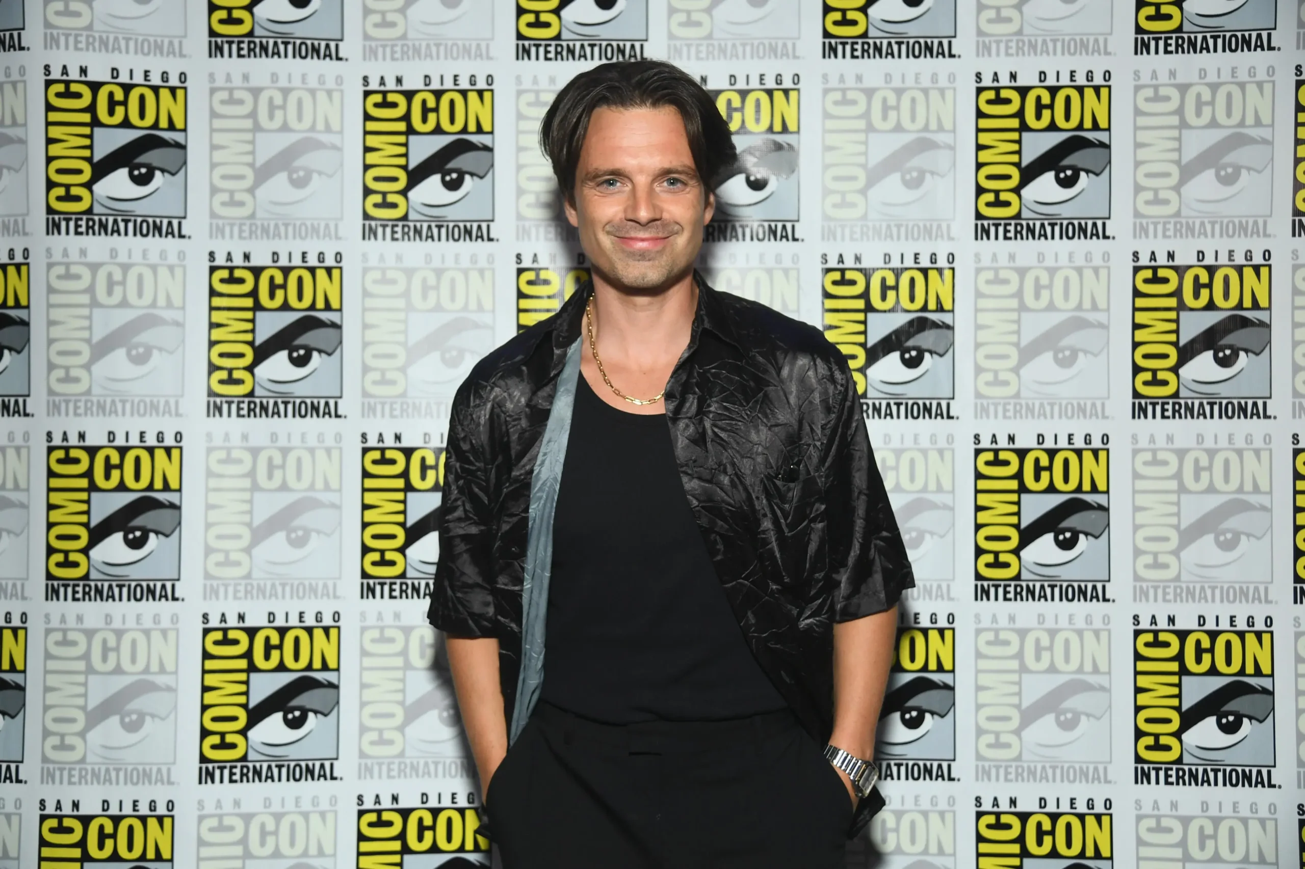 Sebastian Stan smiles while posing at San Diego Comic-Con in front of a repeating Comic-Con International logo wall, wearing a black shirt, textured short-sleeve jacket, gold chain necklace, and wristwatch with his hands in his pockets