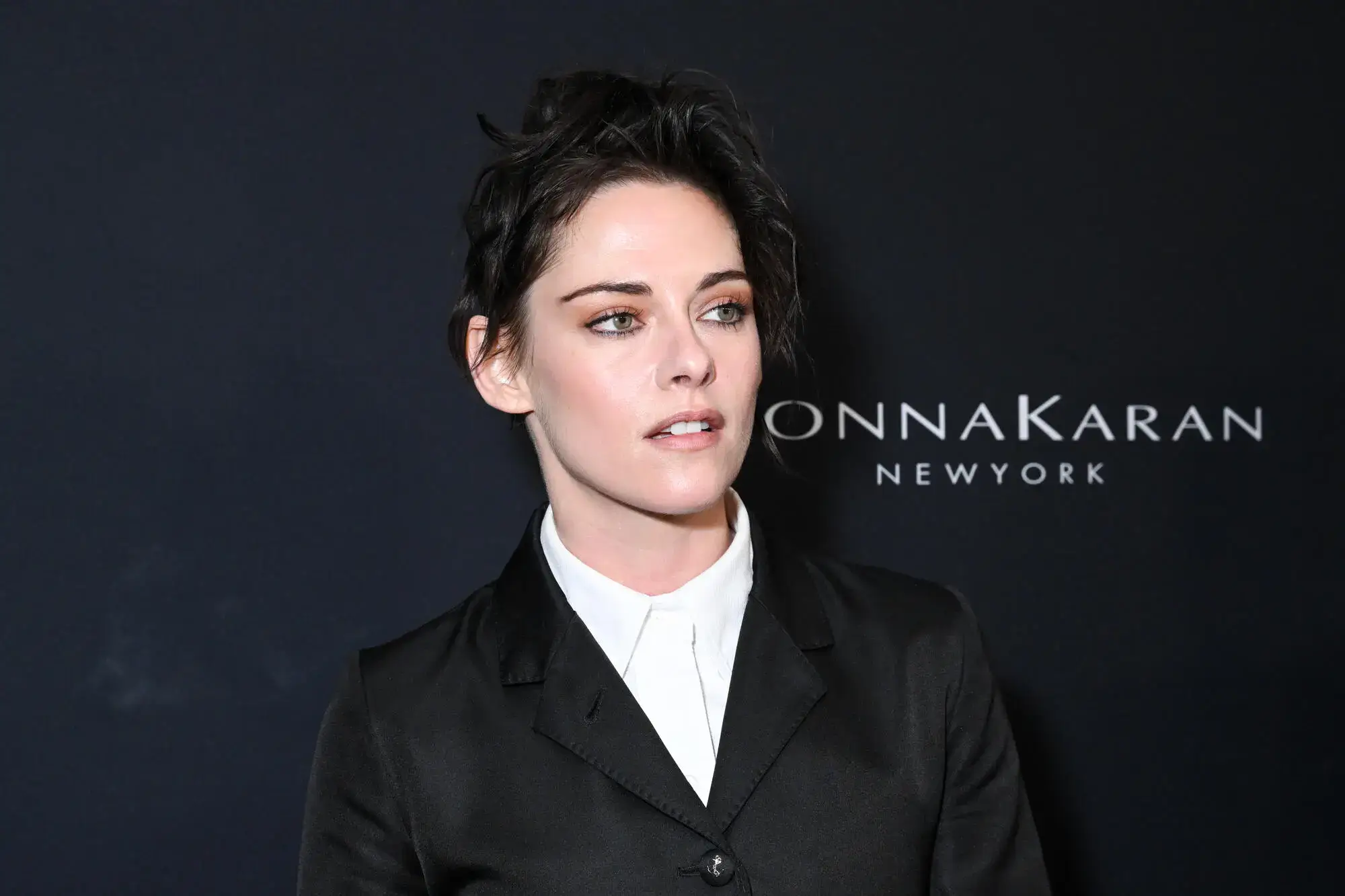 Kristen Stewart wearing a black blazer and white shirt at a red carpet event, posing against a dark backdrop