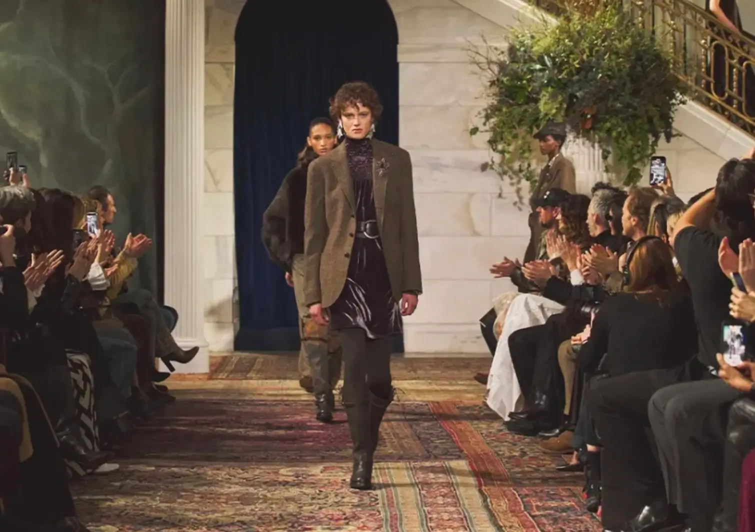 A model walks down a richly carpeted runway during the Ralph Lauren Fall/Winter 2026 show, wearing a tailored brown blazer layered over a deep burgundy velvet look. The setting features marble columns, dark drapery, and lush greenery, while seated guests line both sides of the catwalk, capturing the moment on their phones under warm, moody lighting