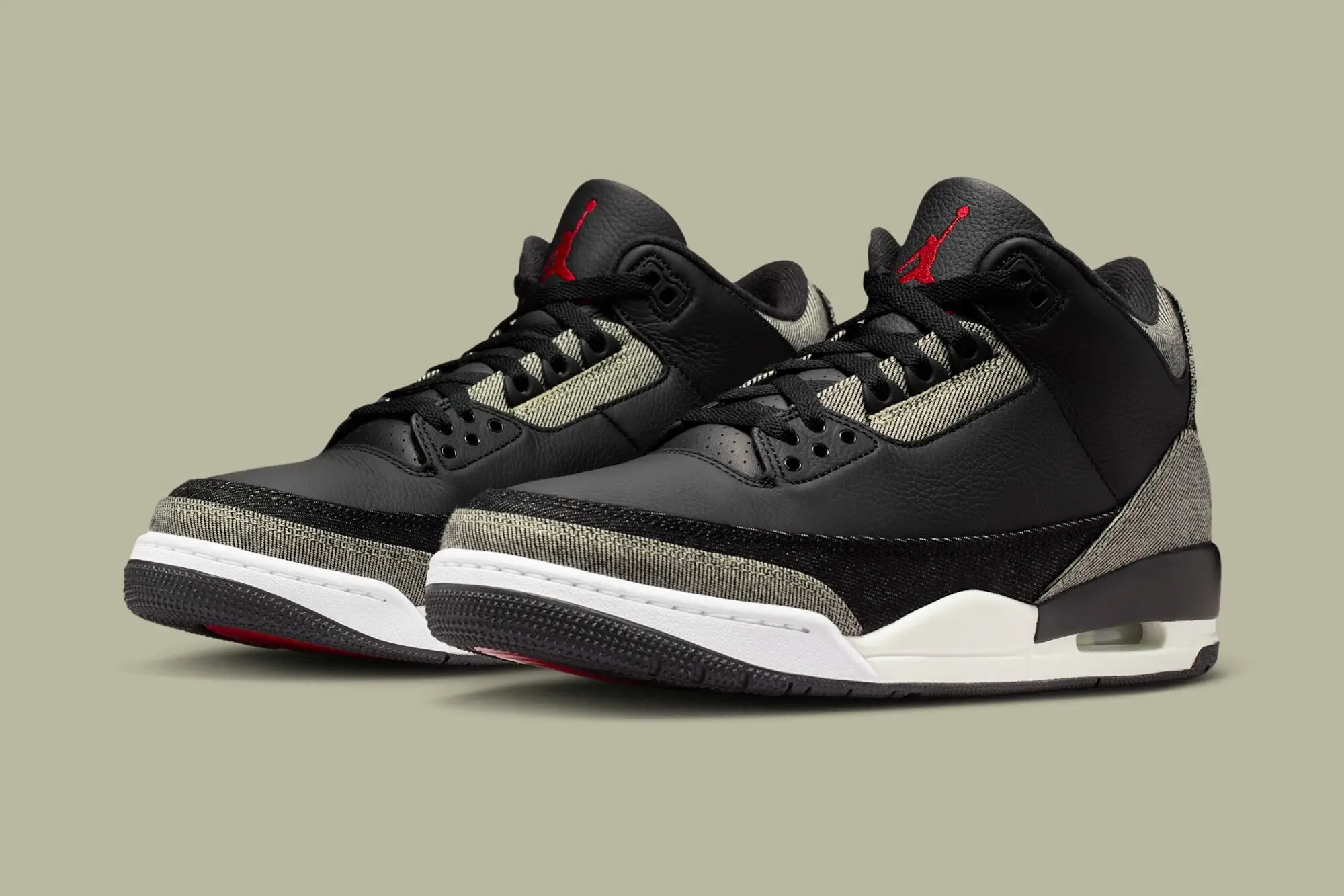 Angled front three-quarter view of the Levi’s x Air Jordan 3 “Black Denim” pair featuring black tumbled leather uppers, contrasting grey and dark denim overlays, red Jumpman logos on the tongues, off-white midsoles with visible Air units, and black rubber outsoles with red traction accents