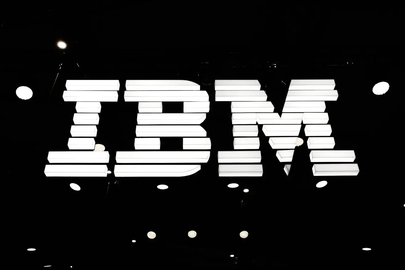White illuminated IBM logo signage suspended against a dark ceiling background, photographed in a high-contrast corporate event or office setting