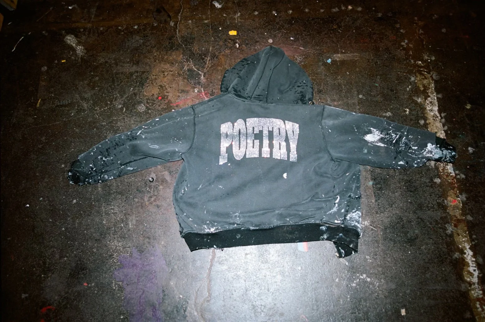 Paint-splattered dark hoodie laid flat on a worn studio floor, featuring bold “POETRY” lettering across the back, with visible distressing and pigment marks suggesting an artist’s working environment