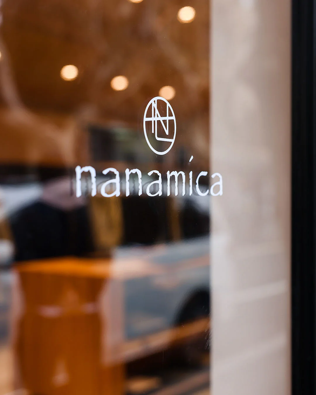 Glass storefront detail featuring the Nanamica logo and wordmark in white, with a softly blurred interior behind it showing warm wood tones, ambient lighting, and indistinct clothing displays that suggest a calm, minimalist retail environment