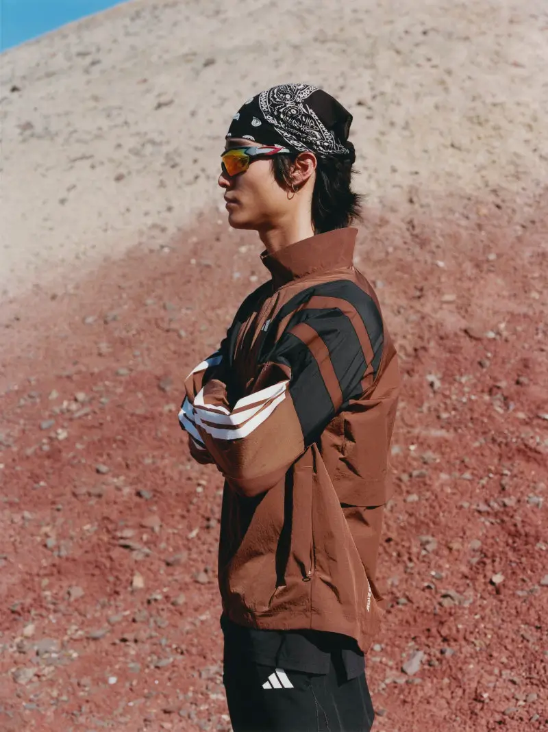 adidas x humanrace performance apparel look featuring a model in a brown lightweight running jacket with black and white stripe detailing, styled with sport sunglasses and bandana, set against a mars-like red and sand-toned landscape