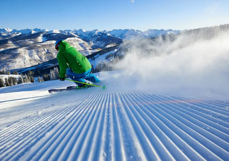 Skier carving across freshly groomed corduroy snow on a wide alpine slope with mountain ranges in the background at a major ski resort operated by Vail Resorts