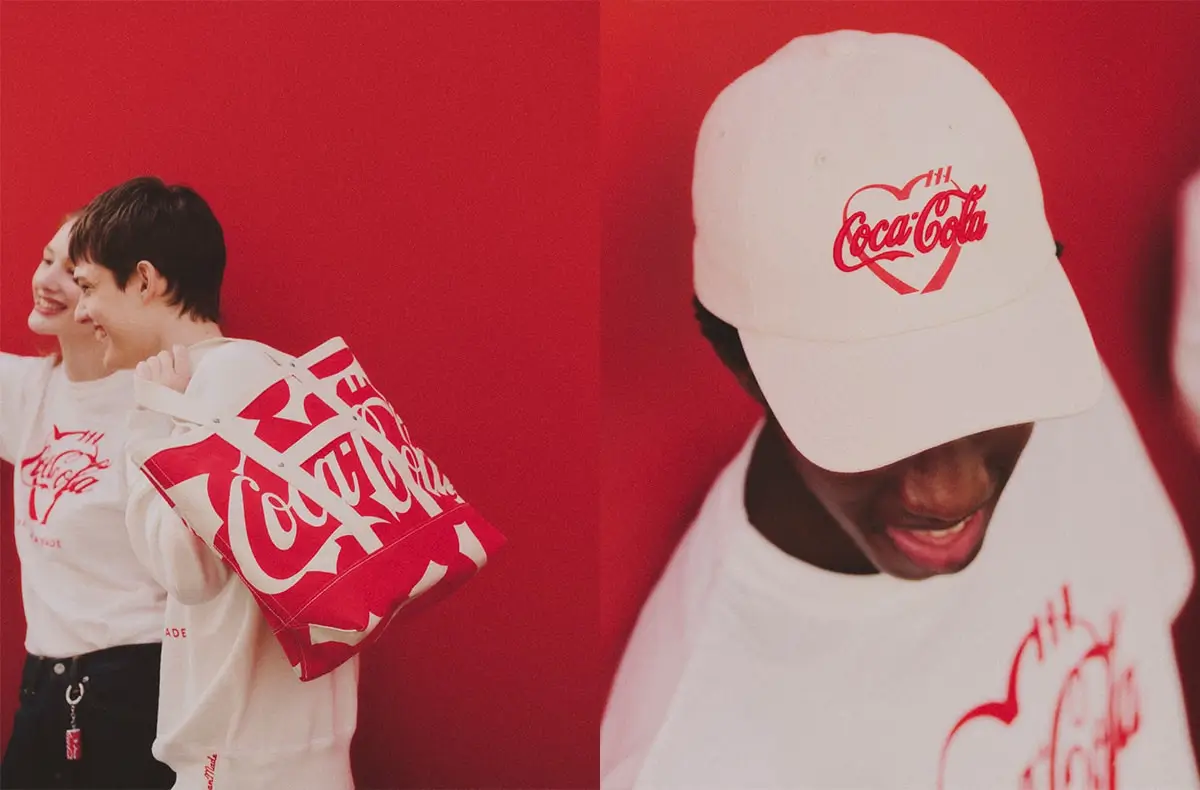 Two models styled against a bold Coca-Cola red backdrop wearing HUMAN MADE collaboration pieces, including a white graphic T-shirt, a red-and-white Coca-Cola logo tote bag, and a white cap featuring a heart-shaped Coca-Cola motif, blending playful streetwear with iconic branding