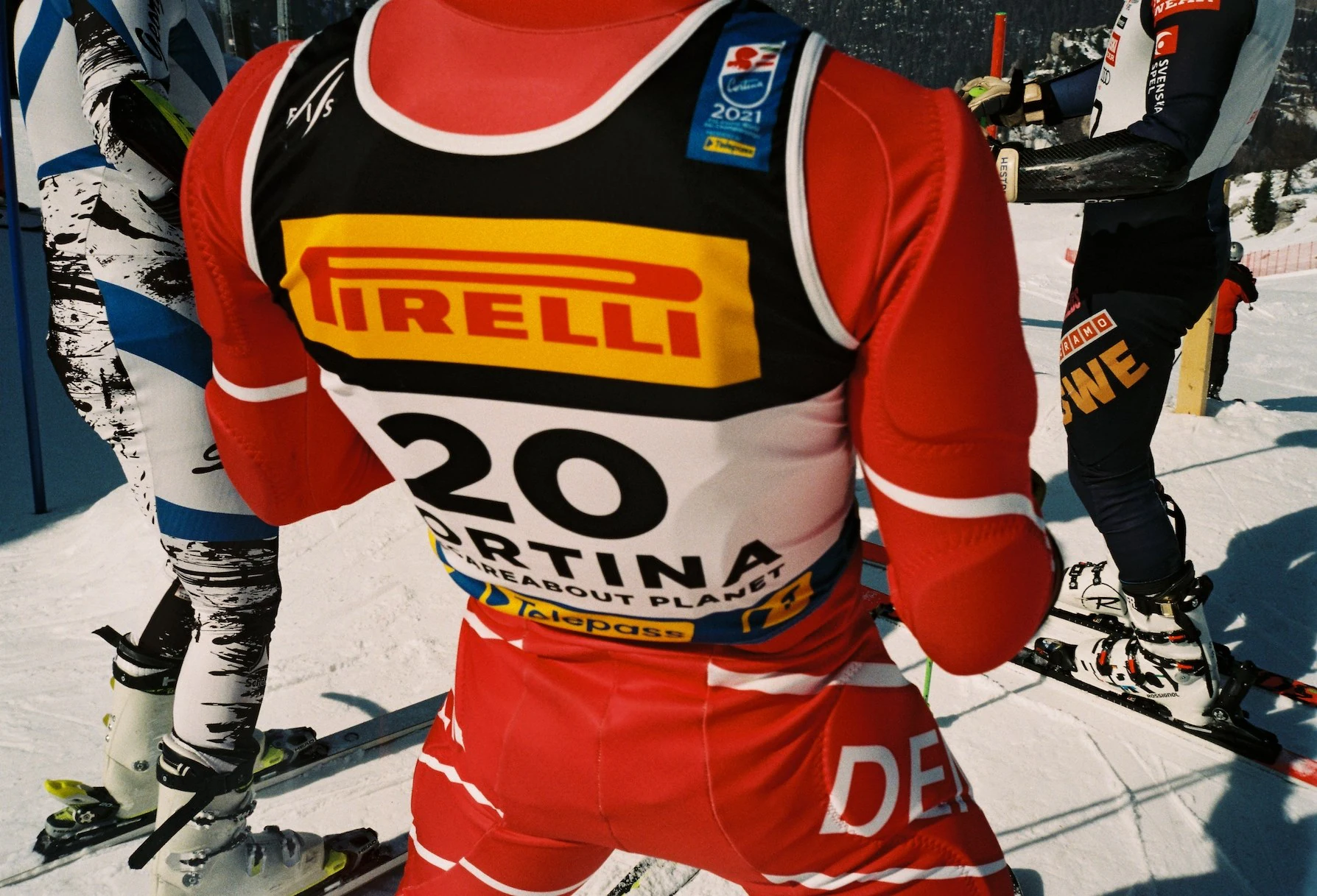 Alpine ski racer wearing a red racing suit and bib number 20 with Pirelli branding at the start gate in Cortina d’Ampezzo during a downhill competition