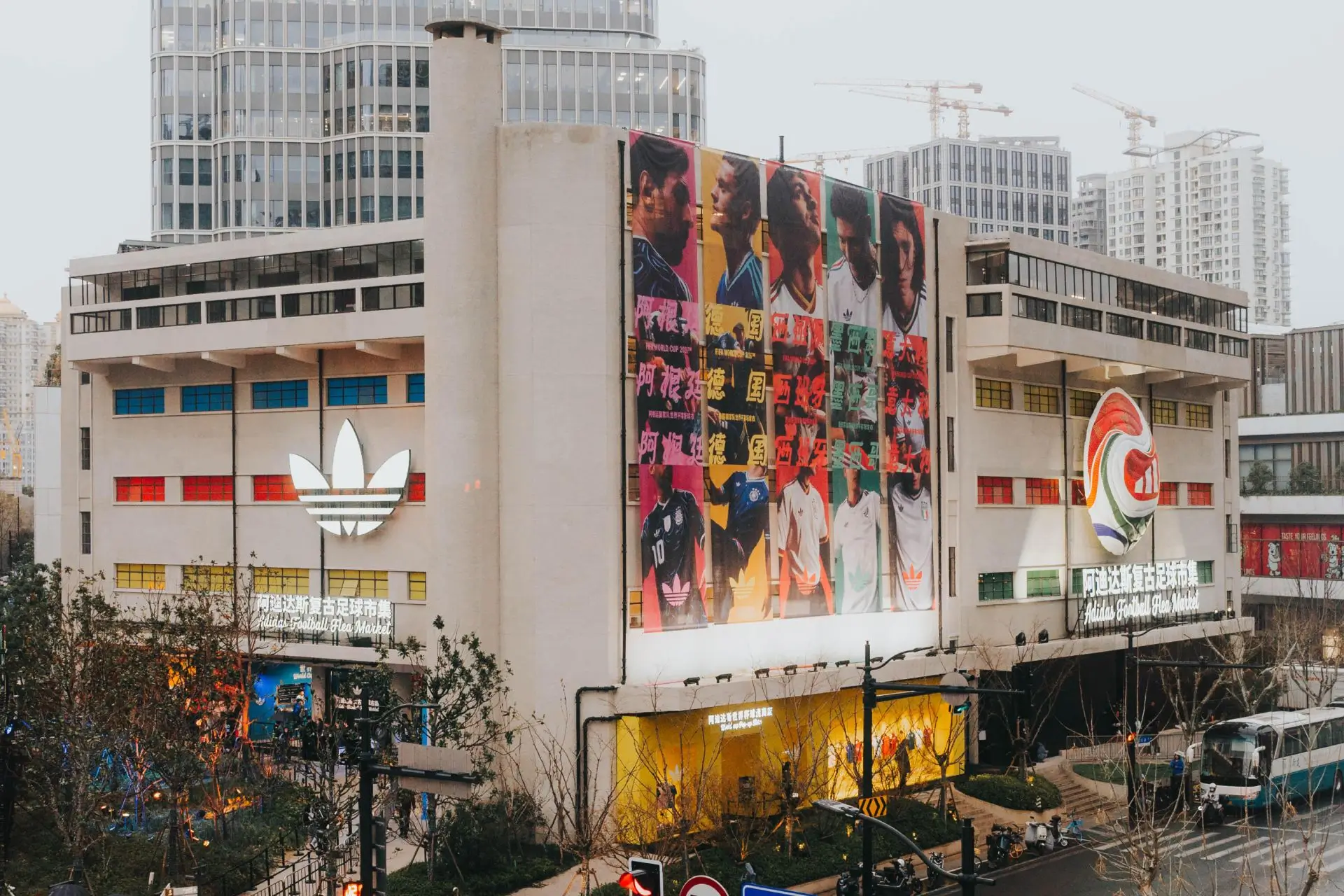 Street-level view of adidas’ Shanghai football flea market experience, showing a large multi-story building wrapped in vibrant football-themed posters, prominent adidas trefoil logos, and an active entrance area connecting the event to the surrounding urban environment