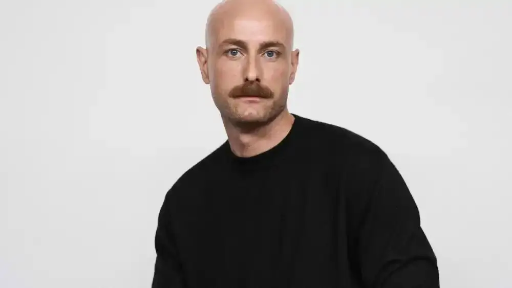 Drew Henry with a neatly groomed mustache wearing a black crewneck sweater, facing slightly to the side against a clean, neutral studio background with soft lighting