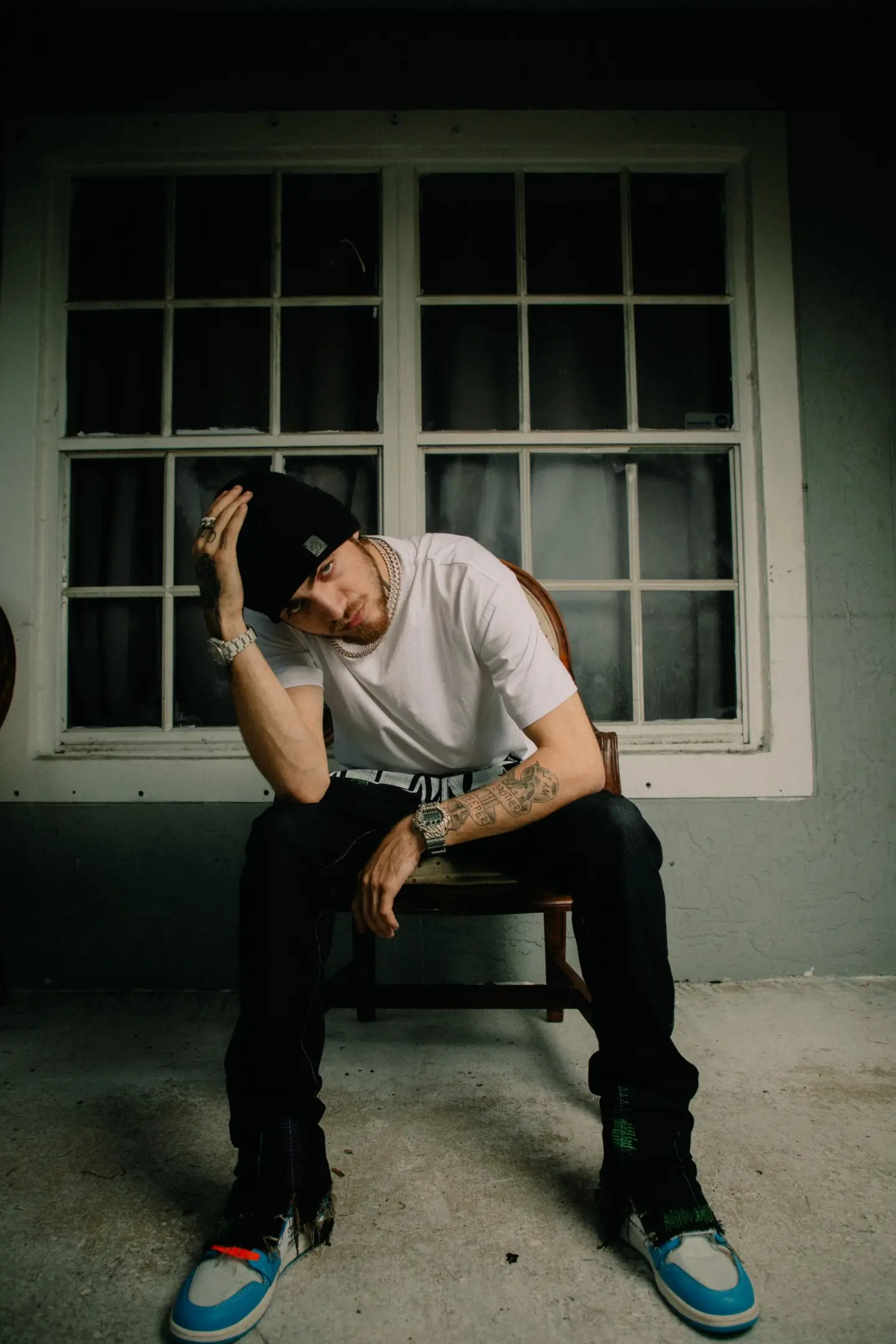 Rapper 1900Rugrat sits on a wooden chair in front of a window at night, wearing a black beanie, white T-shirt, chains, and blue-and-white sneakers while leaning forward with his hand on his head