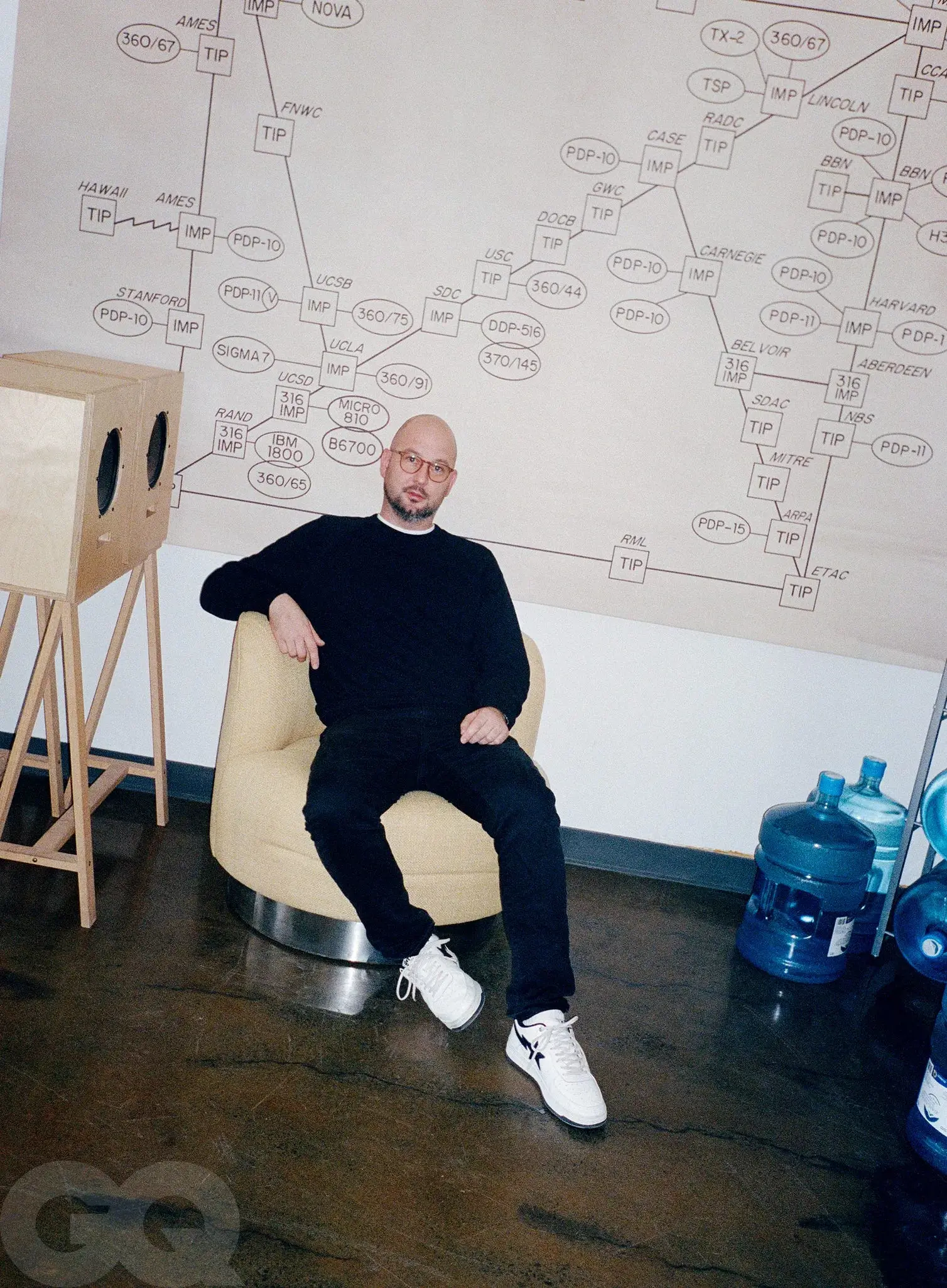 Justin Saunders dressed in a black sweater and black pants sits relaxed in a low, rounded beige chair, wearing white sneakers, in a minimalist studio space. Behind him is a large wall graphic featuring a complex diagram of interconnected nodes and labels, while nearby objects include a wooden speaker-like structure and large blue water jugs on a polished concrete floor