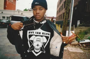 1900Rugrat standing on an urban street, opening his jacket to reveal a graphic skull T-shirt while wearing a black beanie and chain necklace