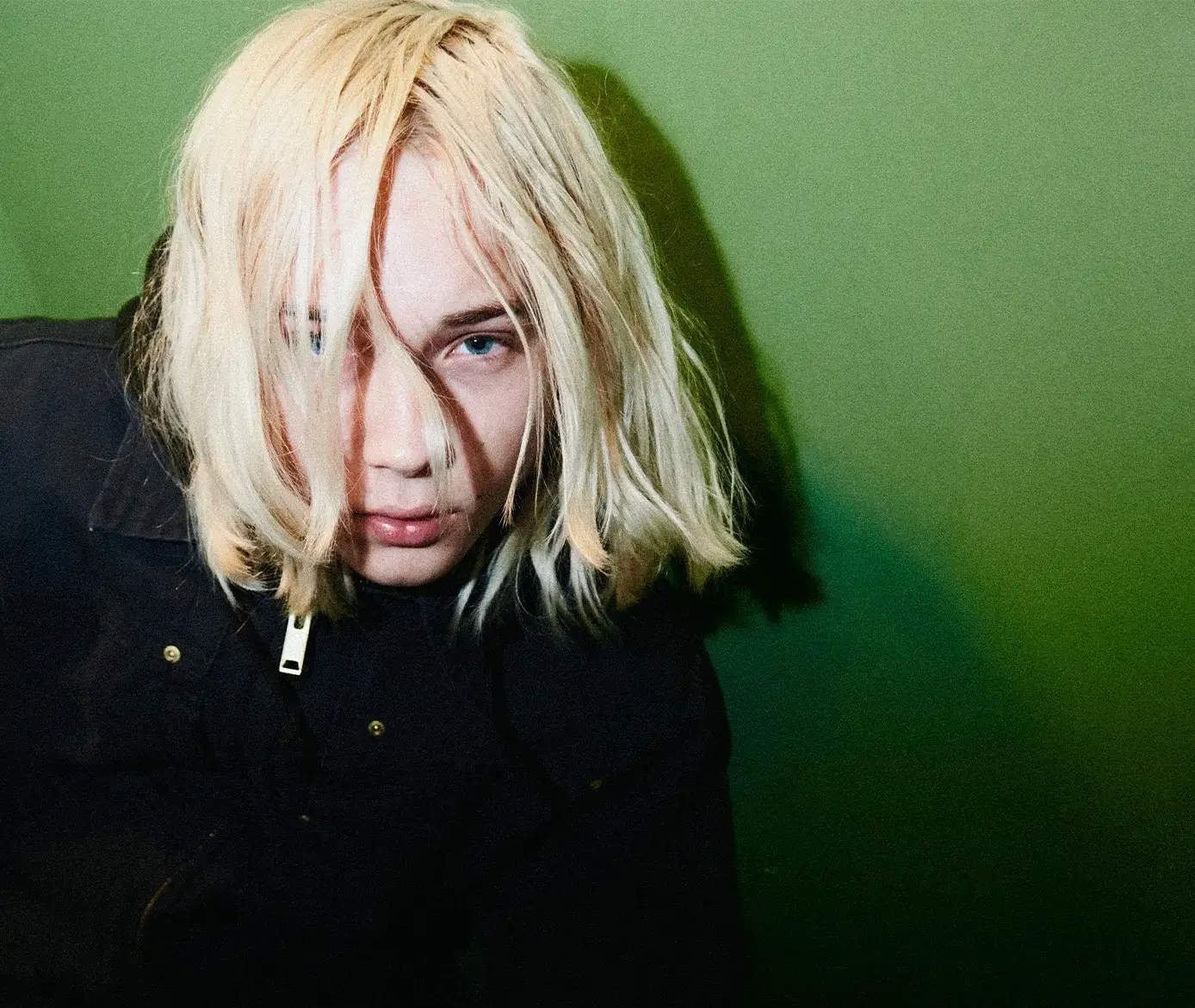 Portrait of Nettspend with long platinum-blonde hair partially covering his face, staring into the camera against a vivid green background while wearing a dark jacket