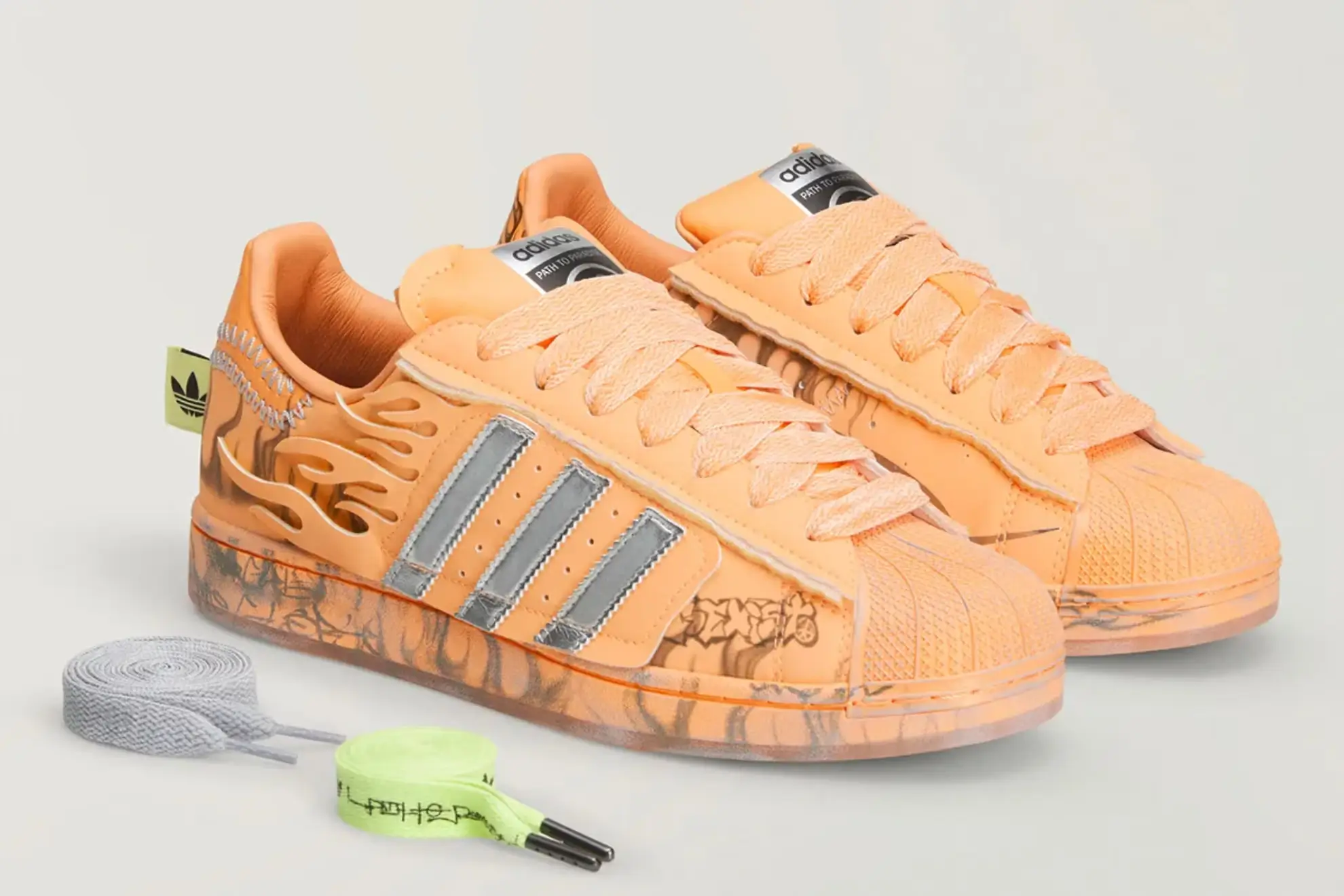 Hellstar x adidas Superstar ‘Hazy Orange’ sneakers featuring a muted peach-orange upper with flame-like cutout detailing, metallic silver Three Stripes, tonal laces, and classic rubber shell toe, paired with alternate grey and neon green lace options against a clean studio background