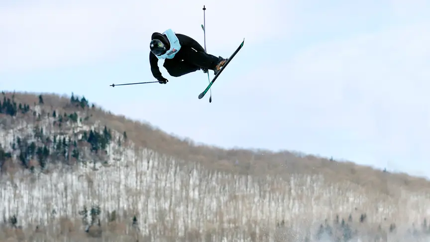 Freeskier mid-air during a big air jump, performing a stylish grab with skis crossed, silhouetted against a pale winter sky with snow-covered mountains in the background