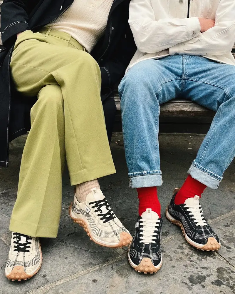 Two people seated on a wooden bench wearing Ami Paris Mirage sneakers—one pair in cream with black stitching and gum studded sole styled with olive green trousers, the other in black with white stitching paired with cuffed denim and bright red socks