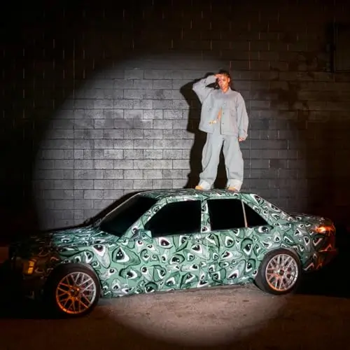 Samara Cyn standing on top of a patterned car under a spotlight at night, wearing a loose grey outfit against a dark brick wall backdrop, in promotional imagery for the song “Oooshxt.”
