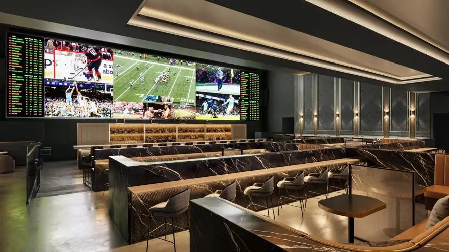 Modern haute sportsbook interior with tiered seating, marble counters, and a massive LED video wall displaying multiple live sports games and betting odds.