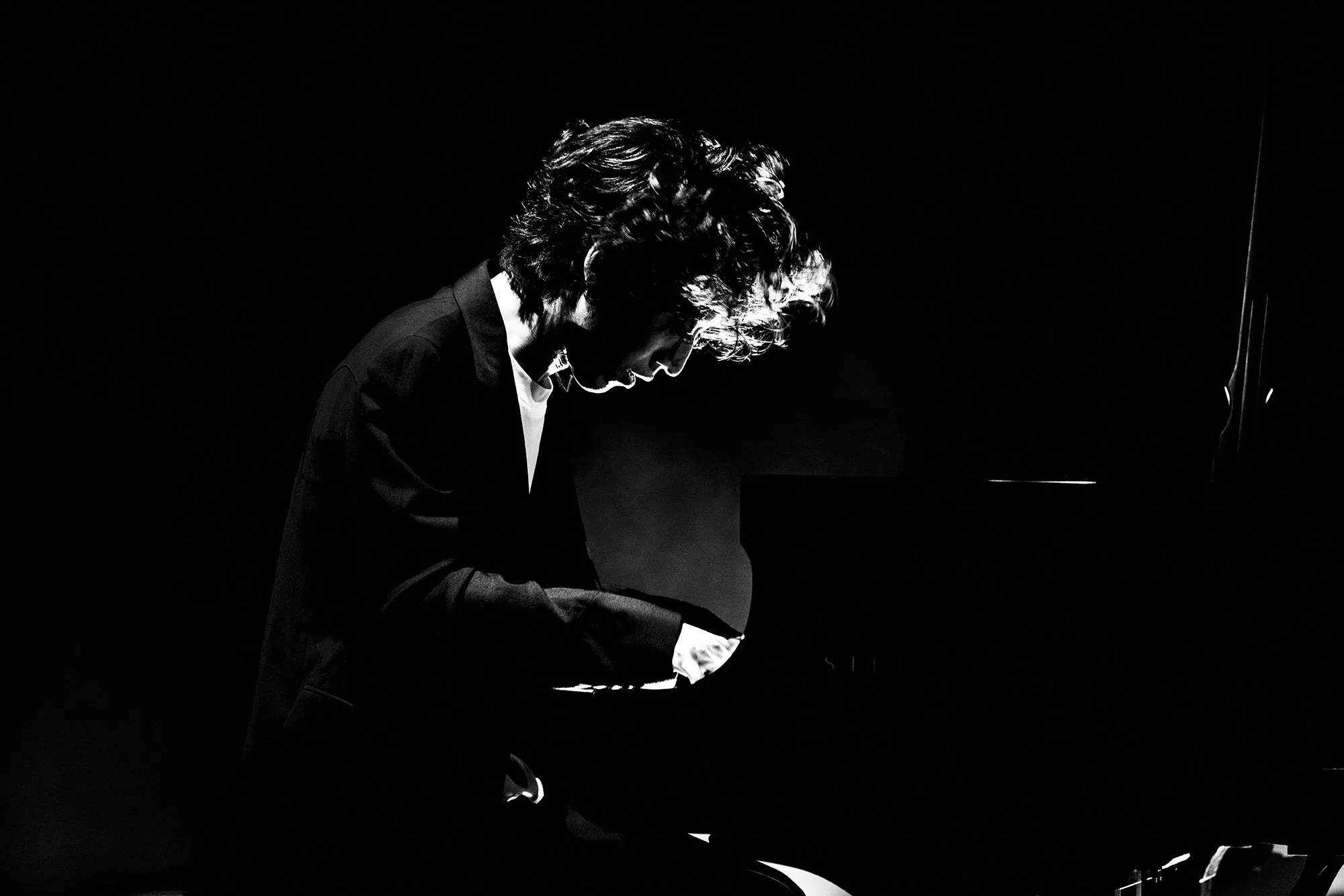 Black-and-white portrait of Hayato Sumino seated at a grand piano, captured in a moment of intense focus, with dramatic lighting emphasizing his silhouette and expressive posture during performance