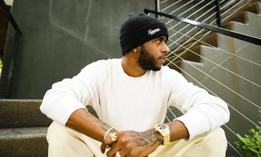 6LACK seated on outdoor steps wearing a black beanie and cream knit outfit, looking to the side with a contemplative expression, showcasing tattoos, gold jewelry, and a watch in a minimalist urban setting