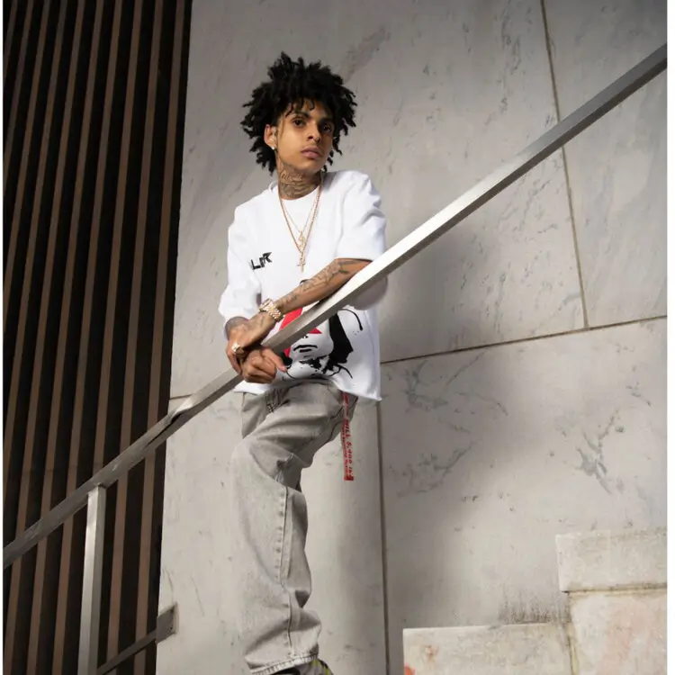 Lil Tony leaning casually on a metal stair railing in a minimalist stone interior, wearing a white graphic t-shirt, light-wash jeans, and gold jewelry, with tattoos visible and a calm, focused expression