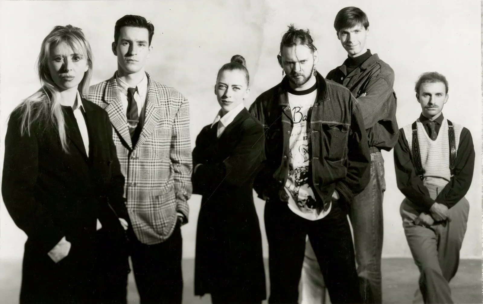 Black-and-white archival portrait of six young designers standing side by side, dressed in a mix of tailored coats, structured suiting, and casual layered pieces. Their expressions are serious and self-assured, capturing an early moment of individuality and collective presence, with styling that reflects late-1980s avant-garde fashion sensibilities