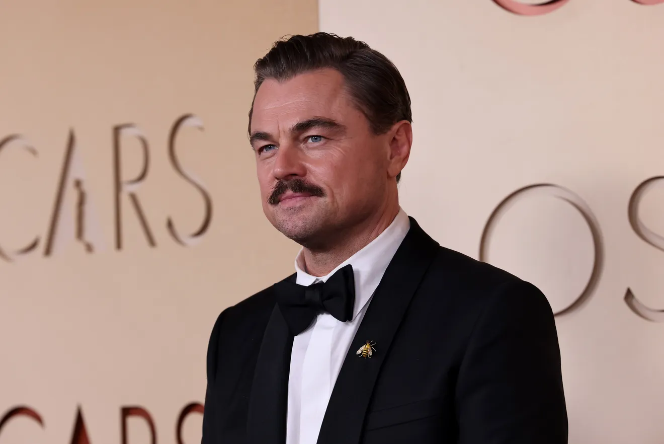 Leonardo DiCaprio wearing a black tuxedo with a bow tie and a small gold bee pin, posing on the Oscars red carpet against a beige backdrop with subtle lettering, his hair slicked back and a trimmed mustache giving a refined, classic look