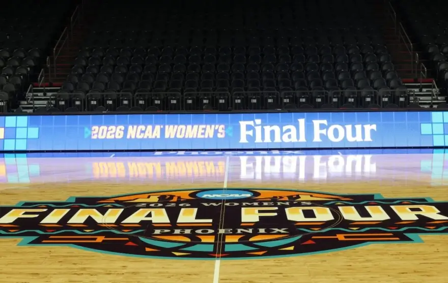 A wide-angle view of an empty basketball arena set for the 2026 NCAA Women’s Final Four in Phoenix. The polished hardwood court prominently features a large, colorful “Final Four Phoenix 2026” center logo, while digital sideline boards display “2026 NCAA Women’s Final Four.” Rows of dark, unoccupied seats rise in the background, emphasizing a pre-game, anticipatory atmosphere