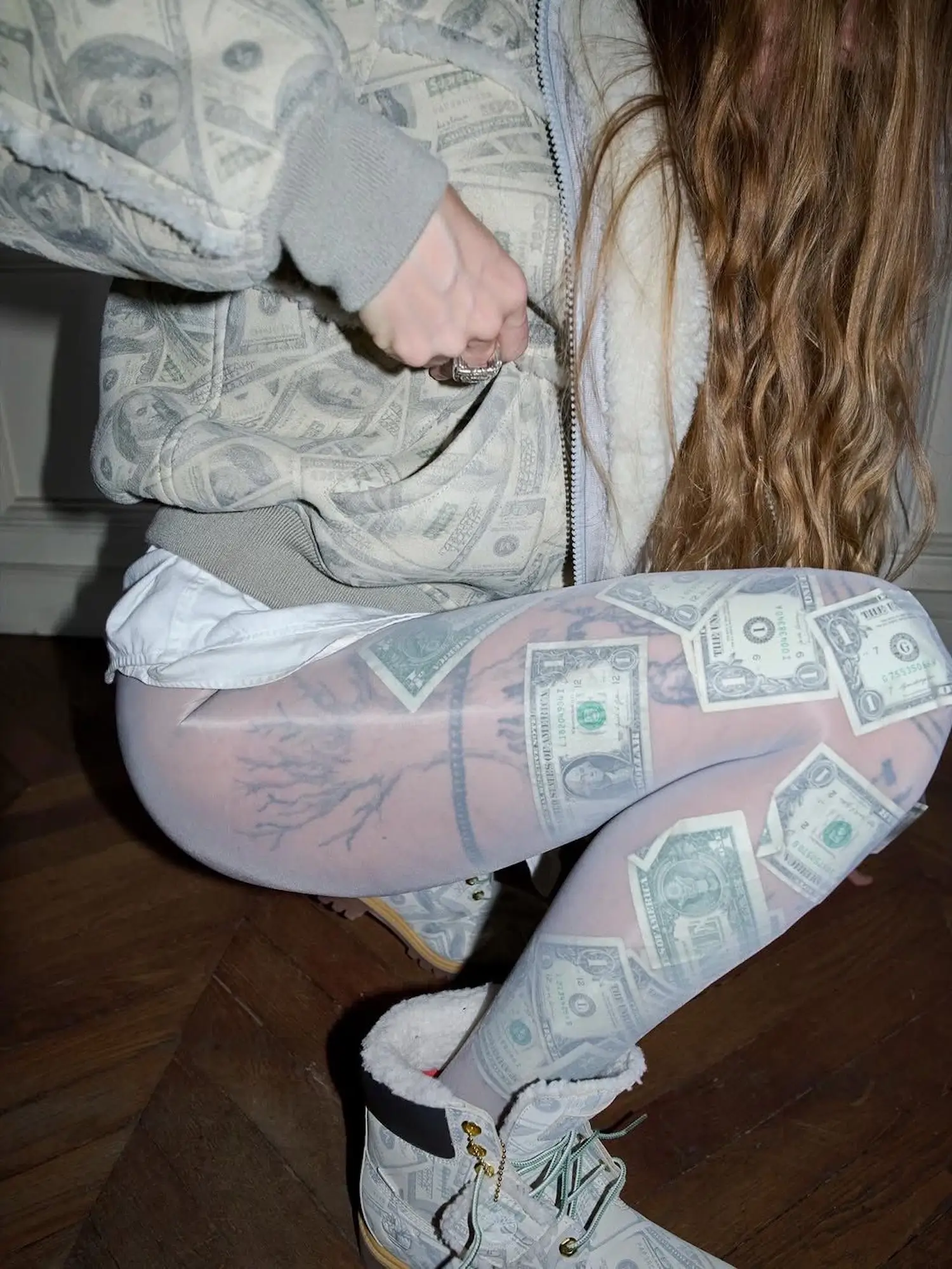 Model wearing Supreme x MM6 Maison Margiela dollar-bill print hoodie and sheer tights layered over tattooed legs, paired with shearling-lined money-print Timberland boots from the Spring 2026