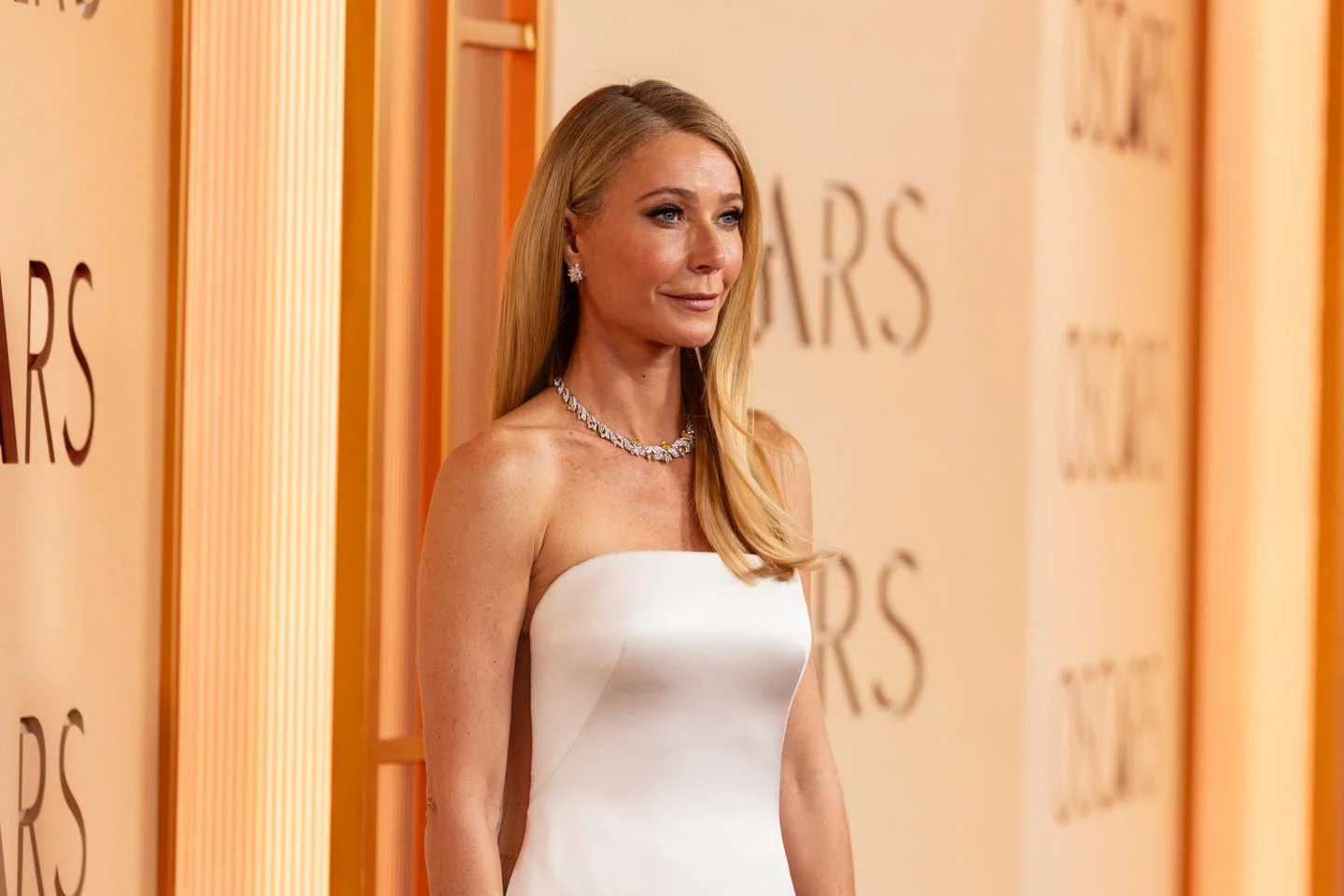 Gwyneth Paltrow on the Oscars red carpet wearing an ivory strapless Giorgio Armani Privé gown, paired with a diamond necklace and earrings, her hair styled straight and side-parted against a warm gold-toned backdrop
